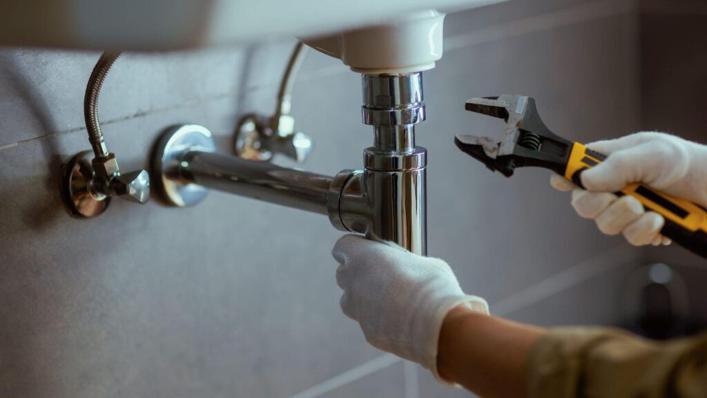 A plumber clearing a drain under a sink with a wrench